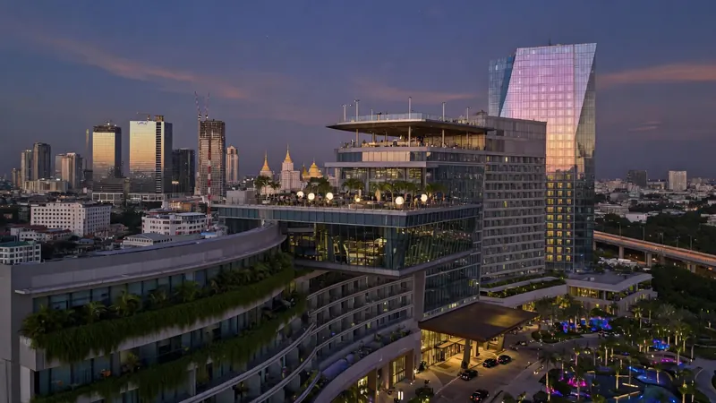Aerial view of a modern cityscape at dusk featuring a large curved hotel with rooftop pool and gardens.